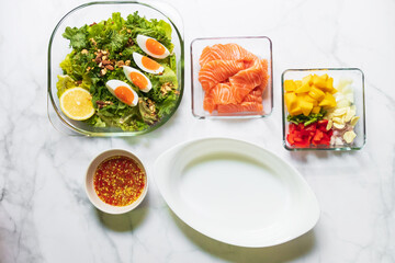 Green salad and kale with boiled egg and oval white bowl with ingredient for delicious spicy salmon mango salad. Top view on white table. 