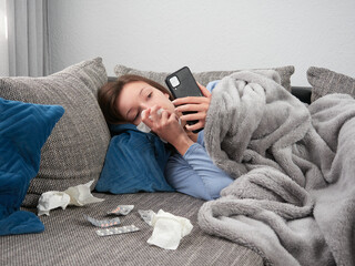 A sick young woman wrapped in a soft blanket is lying on the couch at home. A girl with a runny...