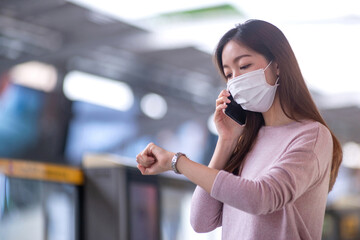 Woman wearing protective mask looks at watch on her wrist at train station platform, waiting...