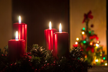 Four red candles burning on advent wreath with blurred colorful christmas tree with glowing lights in the background on evening. Merry Christmas, Advent crown decoration, 4th sunday, Xmas backdrop