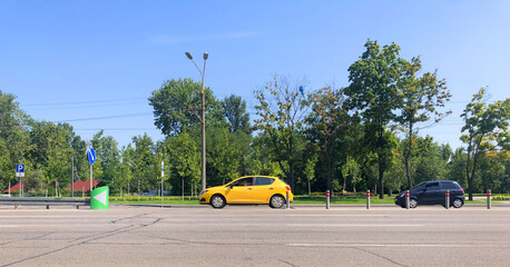 A yellow new car is driving along the road, standing out in the general traffic. A park with green trees, road signs, and a dividing strip with posts In the background. Driving school concept.