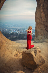 Blonde model girl in a long red dress inside the ring mountain on the background of the city at sunset. On the shoulder is a tattoo of a dream catcher with bird feathers.