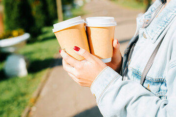 Coffee to go concept. Close-up of female hands with red manicure hold two cardboard disposable cups of coffee outdoors. Selective focus on the right cup