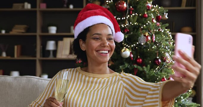 Gen z african american woman wear Xmas cap make series of New Year selfie photos on phone posing on sofa with glass of champagne. Young mixed race lady spend Christmas Eve at home alone shoot stories