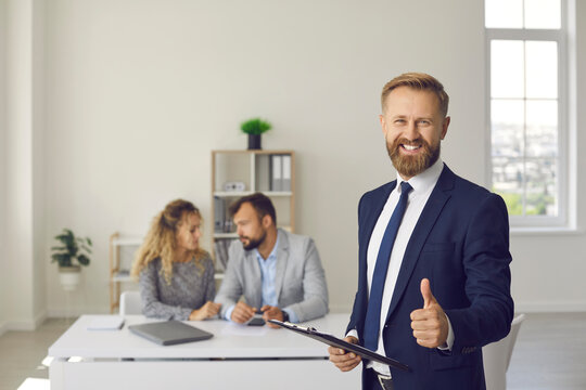 Portrait of happy confident male real estate agent or mortgage broker in suit looking at camera, smiling, offering best deal and giving thumbs up standing in office with his clients in background