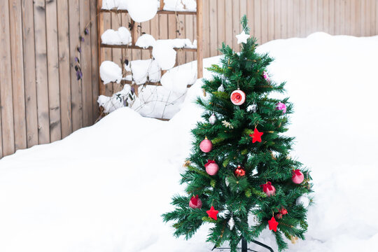 Artificial fir tree decorated with Christmas red toys outdoors in yard in snowy background, New Year and Christmas concept