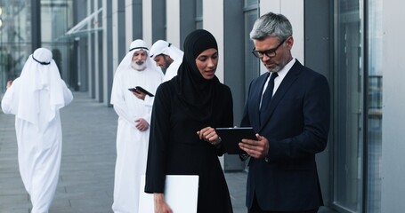 Waist up of multi-ethnic professional managers working near the modern office. Caucasian male manger looking at the tablet screen and talking while muslim woman listening him attentively