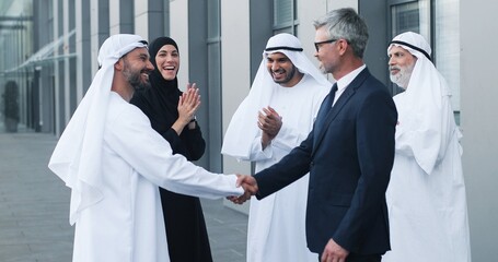 We made a deal. Full length view of successful two business partners shaking hands. They are standing at the street near their colleagues. People are applauded at the background