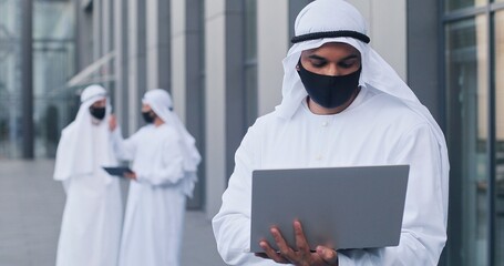 Waist up portrait view of the arabic man wearing protective mask working alone with his laptop...