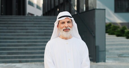 Waist up portrait view of the senior businessman wearing kandura standing at the street at the dinner break and smiling near his office center. Business people concept