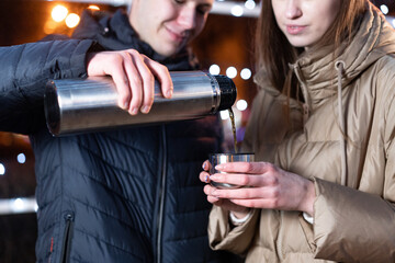 The guy pours tea to the girl in a cup. The girl holds a cup in her hands. Hands in winter.