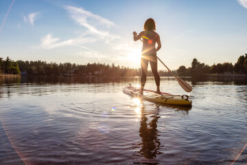 Adventurous Caucasian Adult Woman Paddling on a Stand up Paddle Board in water at a city park. Sunny Sunset Sky. Gorge Park, Victoria, Vancouver Island, BC, Canada. - obrazy, fototapety, plakaty