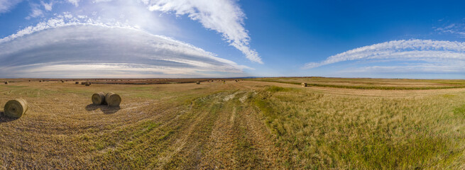 Panorama of flat prairie fields with round hay bales and autumn colored grass. The blue sky has arcing white clouds.
