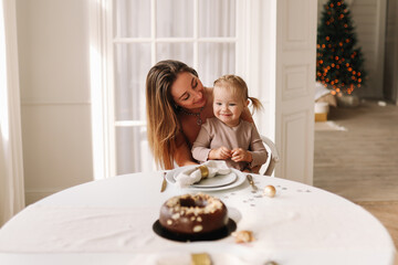 The concept of Christmas. Happy cute mom and daughter a small child are laughing sitting at the dining table celebrating the New Year and Christmas holiday in a cozy room at home on the weekend