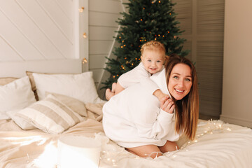 Obraz na płótnie Canvas Smiling, having fun mom and daughter child spend time together playing on the bed by the Christmas tree on a holiday day in a cozy bedroom at home on the weekend. Selective focus