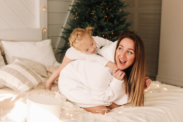 Obraz na płótnie Canvas Smiling, having fun mom and daughter child spend time together playing on the bed by the Christmas tree on a holiday day in a cozy bedroom at home on the weekend. Selective focus