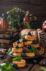many waffles are stacked on a plate, decorated with mint leaves and blueberries. Vegetables are lying around on a wooden table
