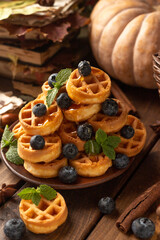 many waffles are stacked on a plate, decorated with mint leaves and blueberries. Vegetables are lying around on a wooden table