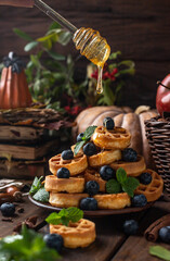 many waffles are stacked on a plate, decorated with mint leaves and blueberries. On top of them, honey pours from glass spoons