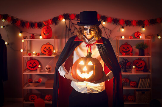 Halloween Man With Pumpkin In Darkness. Portrait Of Man Dressed As Vampire Standing In Room With Red Light With Spooky Smile And Glowing Jack-o-lantern In His Hands. Mystery And Horror Concept.