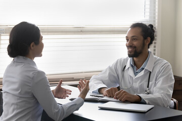 Happy optimistic young doctor listening to patient at appointment in office, talking to Indian woman, smiling. Mixed race millennial practitioner, therapist giving medical consultation, advice
