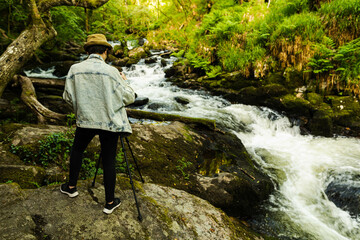 Attractive young female wearing a hat and denim jacket and taking a photo of a river that flows...