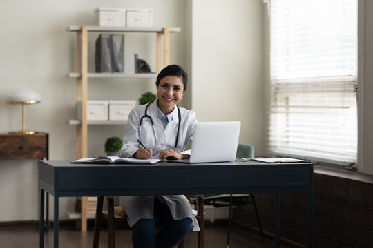 Happy young Indian doctor, therapist, general practitioner at work table with laptop in office, looking at camera, smiling, writing notes, medical records. Medic care concept. Professional portrait
