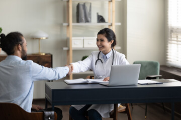 Obraz premium Happy young Indian GP doctor shaking hands with male patient, celebrating successful therapy result. Mixed raced millennial man visiting practitioner, shaking hands, expressing gratitude, respect
