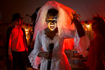 Portrait of black woman in white bridal veil at Halloween horror party at home. Escape room actress who plays Corpse Bride dead girl holding flashlight that lights up face with ugly grimace expression - obrazy, fototapety, plakaty