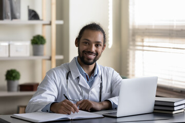 Fototapeta na wymiar Happy Afro American doctor, general practitioner, physician working in hospital office, writing medical records at laptop, looking at camera, smiling. Healthcare service concept. Head shot portrait