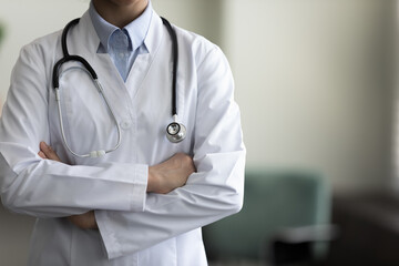Cropped shot of female GP doctor with stethoscope wearing white coat, standing indoors with folded...