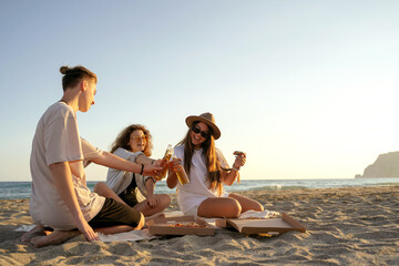 Young happy friends doing picnic seaside. Group of people sitting on the beach, eating pizza and drinking beer together. Summer spending time outdoor concept.