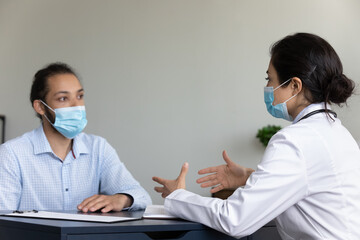 Young Indian doctor and Black mixed raced covid patient wearing face masks, talking at appointment,...