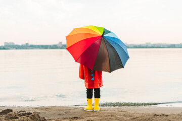 Boy in a red raincoat and yellow rubber boots holds rainbow umbrella standing at beach. Child with...