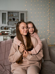 Fototapeta premium a little daughter in a sweatshirt is sitting on the couch and hugging her mother