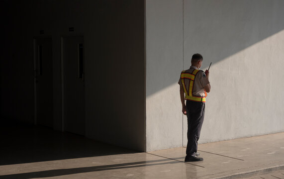 Asian security guard in safety vest using walkie-talkie while working on sidewalk in underground of parking garage