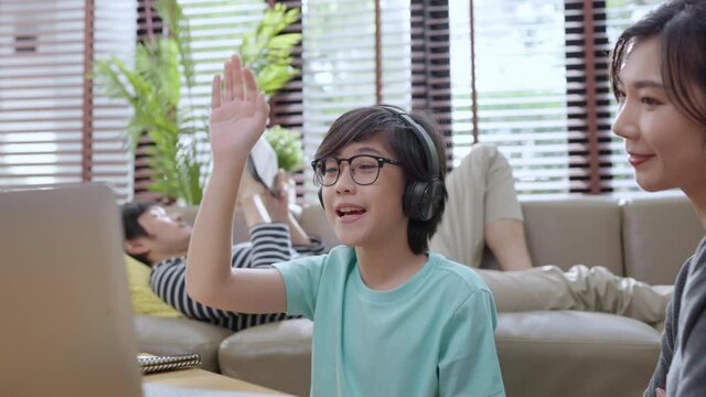 cheerful asian child boy online lesson study from home concept,asian child hand up for answer question from online teacher ,asian boy sit with mom and dad learn from laptop at living area at home