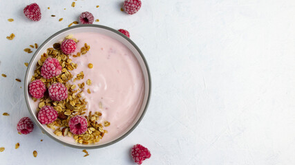 Top view of raspberry yogurt in a white bowl with granola and frozen raspberries on a white plate with a copy space