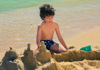 funny curly boy in swimming trunks created a huge castle on the sandy beach