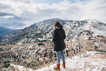 Back view of young woman standing at mountain top observation deck over Vouraikos gorge. Mega Spileon Monastery terrace with splendid snow-covered mountains view. Winter vacation. Greece.