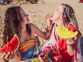 close up portrait of two happy girls eating watermelon at the beach outdoors in summer time. friends having fun and enjoying and biting fresh fruits on seaside holidays.