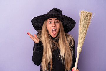 Young Russian woman disguised as a witch holding a broom isolated on purple background surprised and shocked.