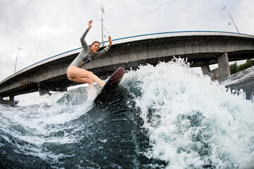 Naklejka premium energetic woman rides down the wave on wakesurf board. Bridge at background