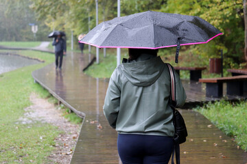 Rain in a city, woman with black umbrella walking in autumn park on people background. Rainy weather, heavy rainfall
