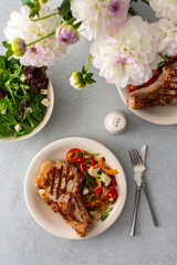 Delicious hearty dinner in plates on the table and a bouquet of beautiful autumn flowers, fried pork entrecote on the bone with stewed vegetables and fresh salad with mozzarella