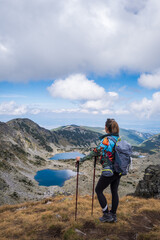 Girl mountain hiker in colorful clothes and with walking sticks standing on a cliff and looking at...