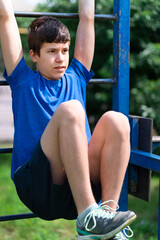 Fototapeta na wymiar teenage boy exercising outdoors, sports ground in the yard, he hangs on the horizontal bar, raises his legs and pumps his abs, healthy lifestyle