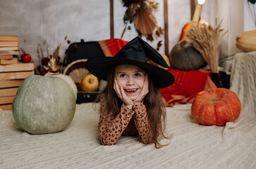a laughing little girl in a witch's hat lies on a knitted blanket with pumpkins