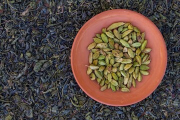 clay bowl full of green cardamoms placed on background bull of dry green tea leaves