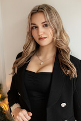 Cute beautiful young woman in fashionable black clothes with an elegant blazer and big breasts stands near a white wall